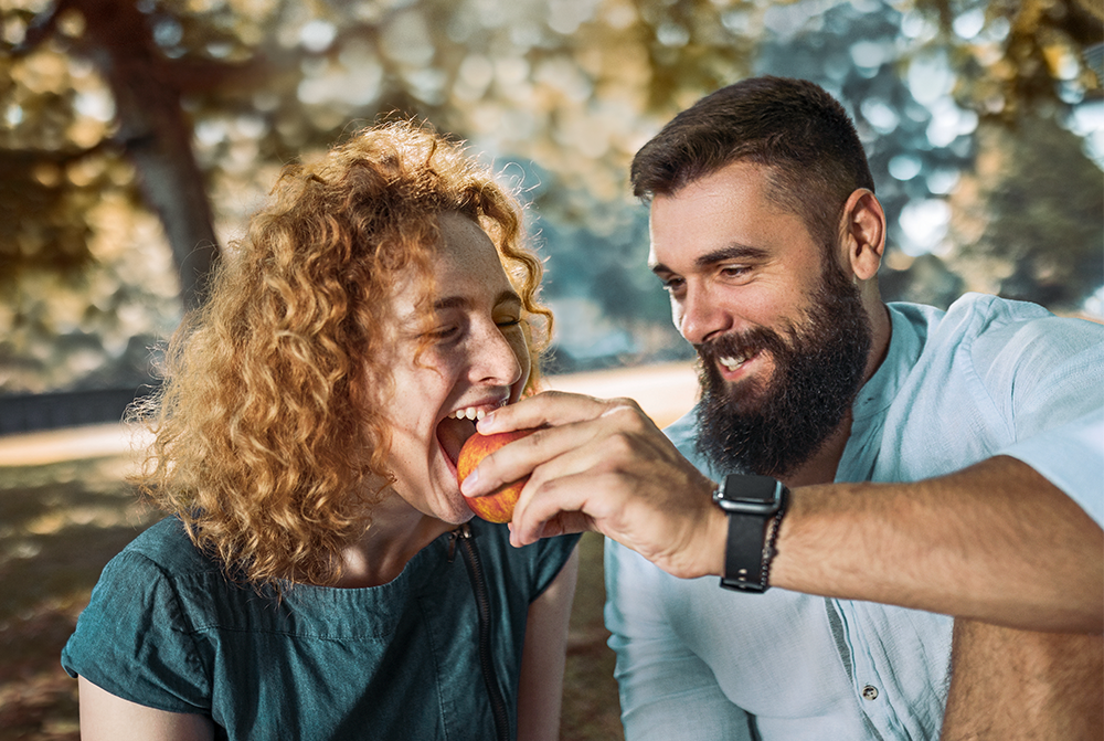 Mann der eine Frau mit einem Apfel füttert, man erkennt strahlende Zähne